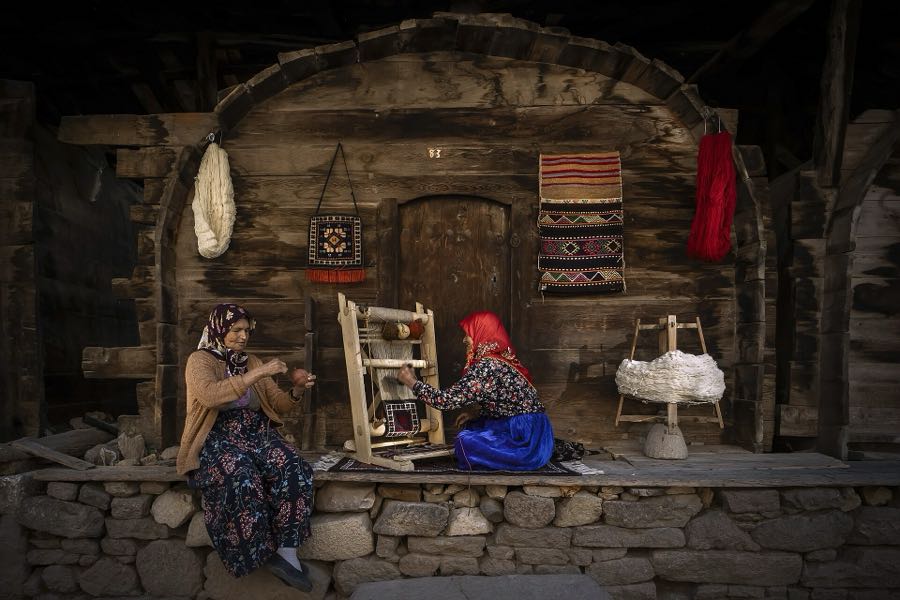 Ahşap bir evin önünde tezgâhta halı dokuyan iki kadın